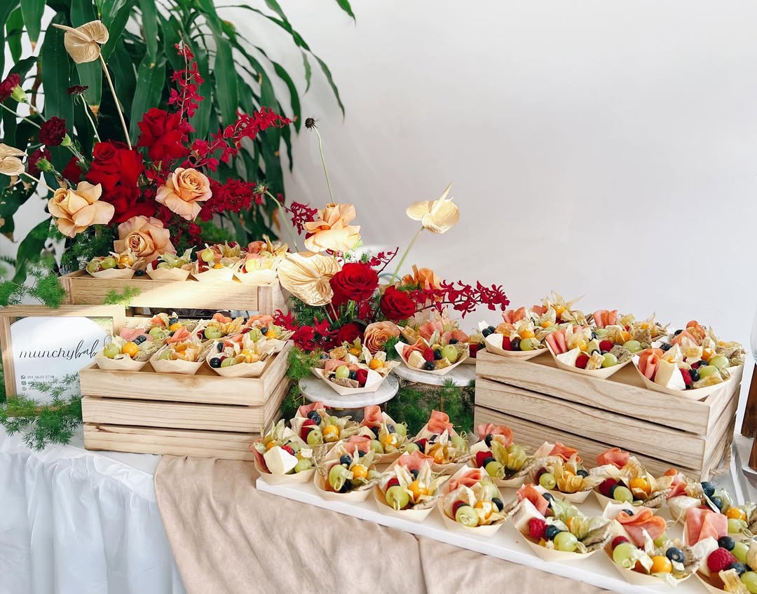 A display of assorted fruit and charcuterie cups arranged in wooden crates and trays, with decorative red and peach flowers in the background—perfect inspiration for pretty dessert tables and memorable dessert displays.