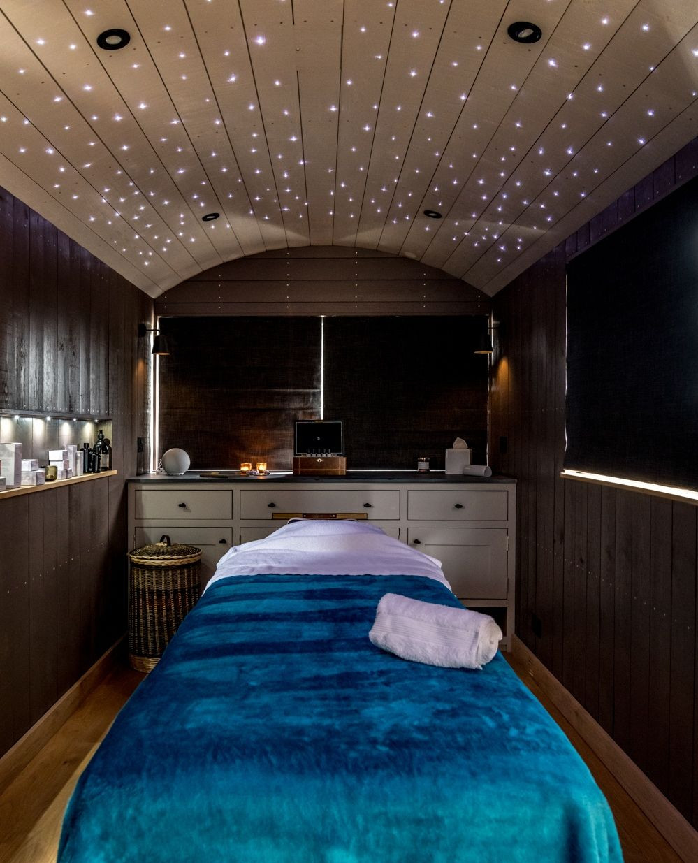 A massage room with a blue-covered treatment table, a rolled towel, and shelves with products showcases soothing massage room décor, with dark walls and a ceiling of tiny star-like lights, creating the perfect relaxation space.