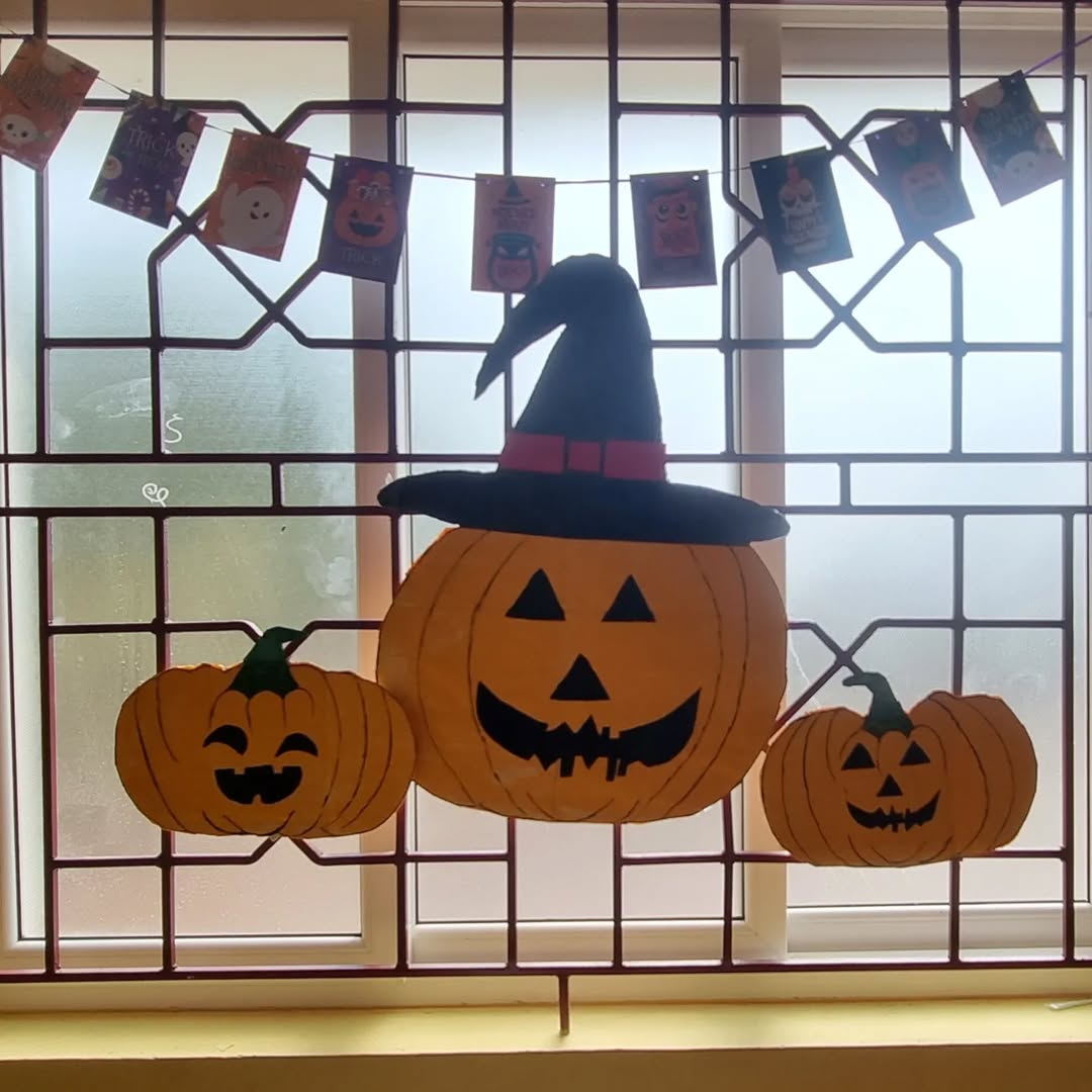Three paper jack-o’-lanterns, one wearing a witch hat, are displayed on a window with a Halloween-themed banner above—festive Halloween window decorations perfect for spooky season flair.