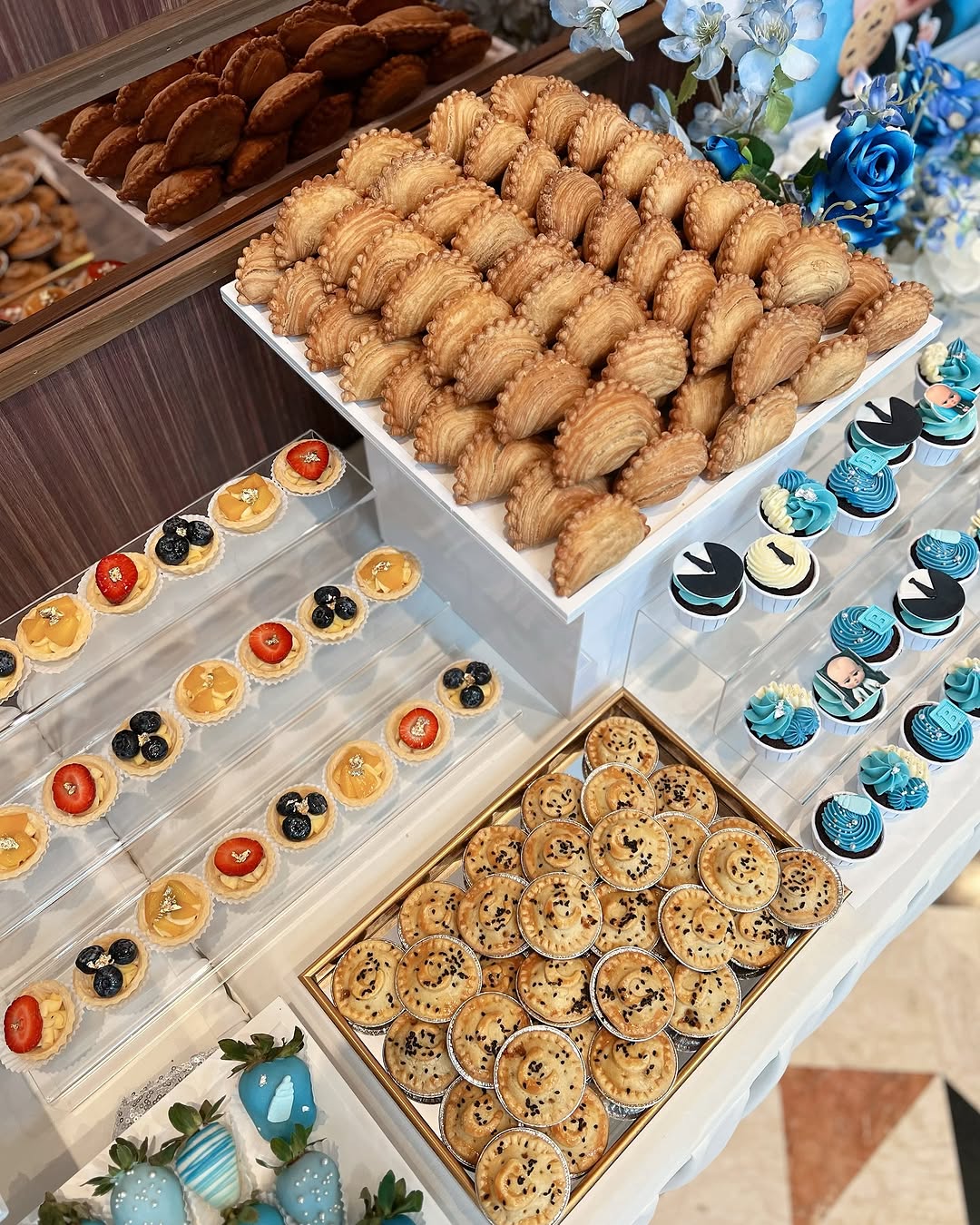 A dessert table perfect for occasion desserts, featuring assorted pastries—filled cookies, fruit tarts, cupcakes, cheesecakes with fresh fruit, and blue-themed treats—neatly arranged on stands for inspired dessert table ideas.