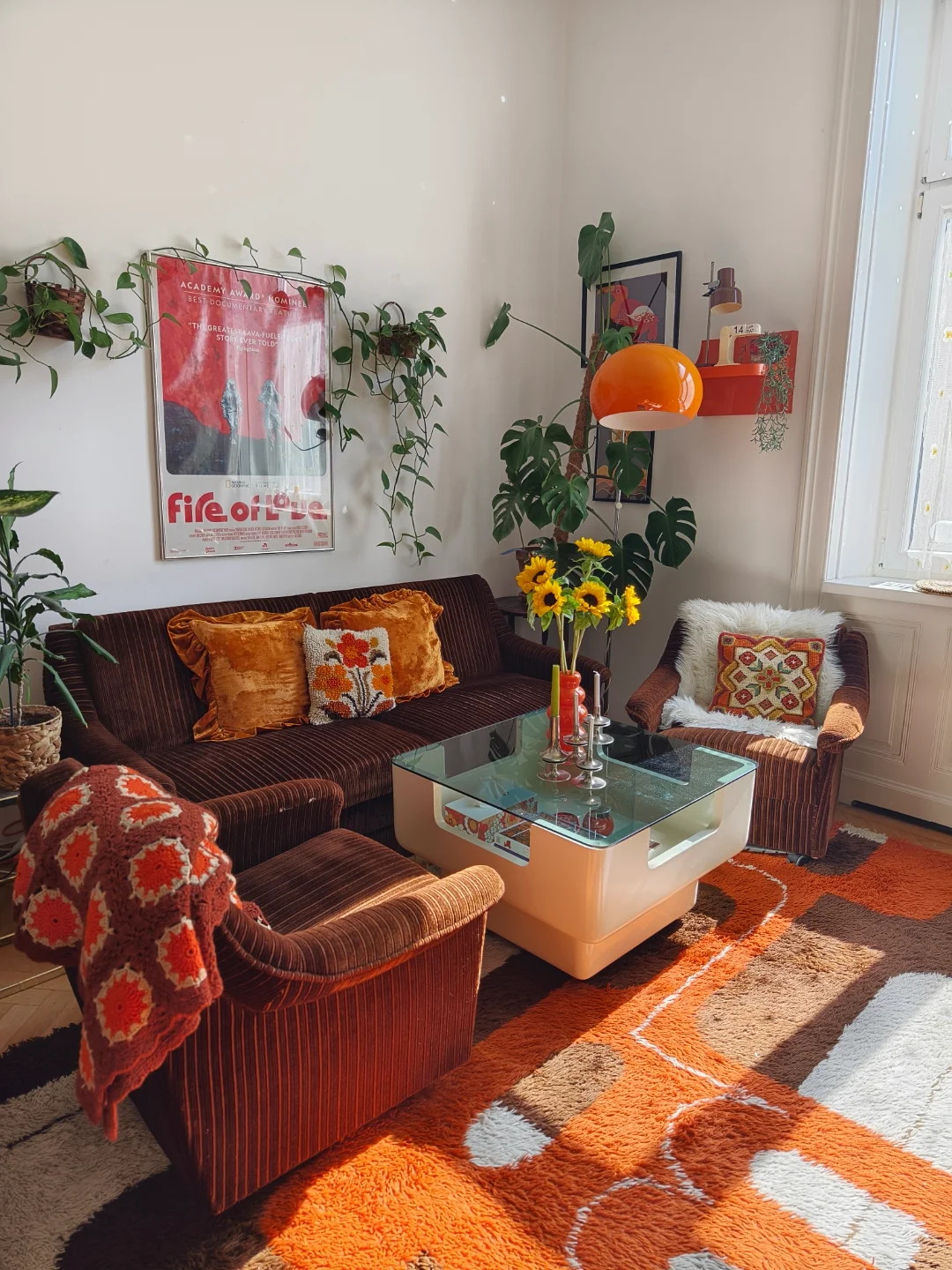 A retro-style living room with chic dark brown sofas, orange furniture, a glass coffee table, sunflowers in a vase, and wall art, bathed in sunlight streaming through the window—perfect for inspiring new living room ideas.