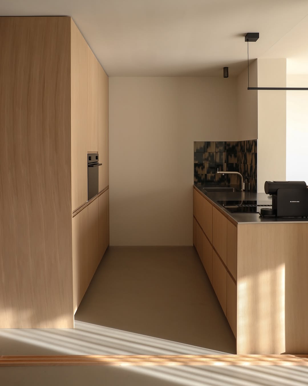 Minimalist oak kitchen with light wood cabinets, built-in oven, black countertop, geometric tile backsplash, and a coffee machine on the counter. Natural light casts shadows on the floor—perfect for modern kitchen design enthusiasts.