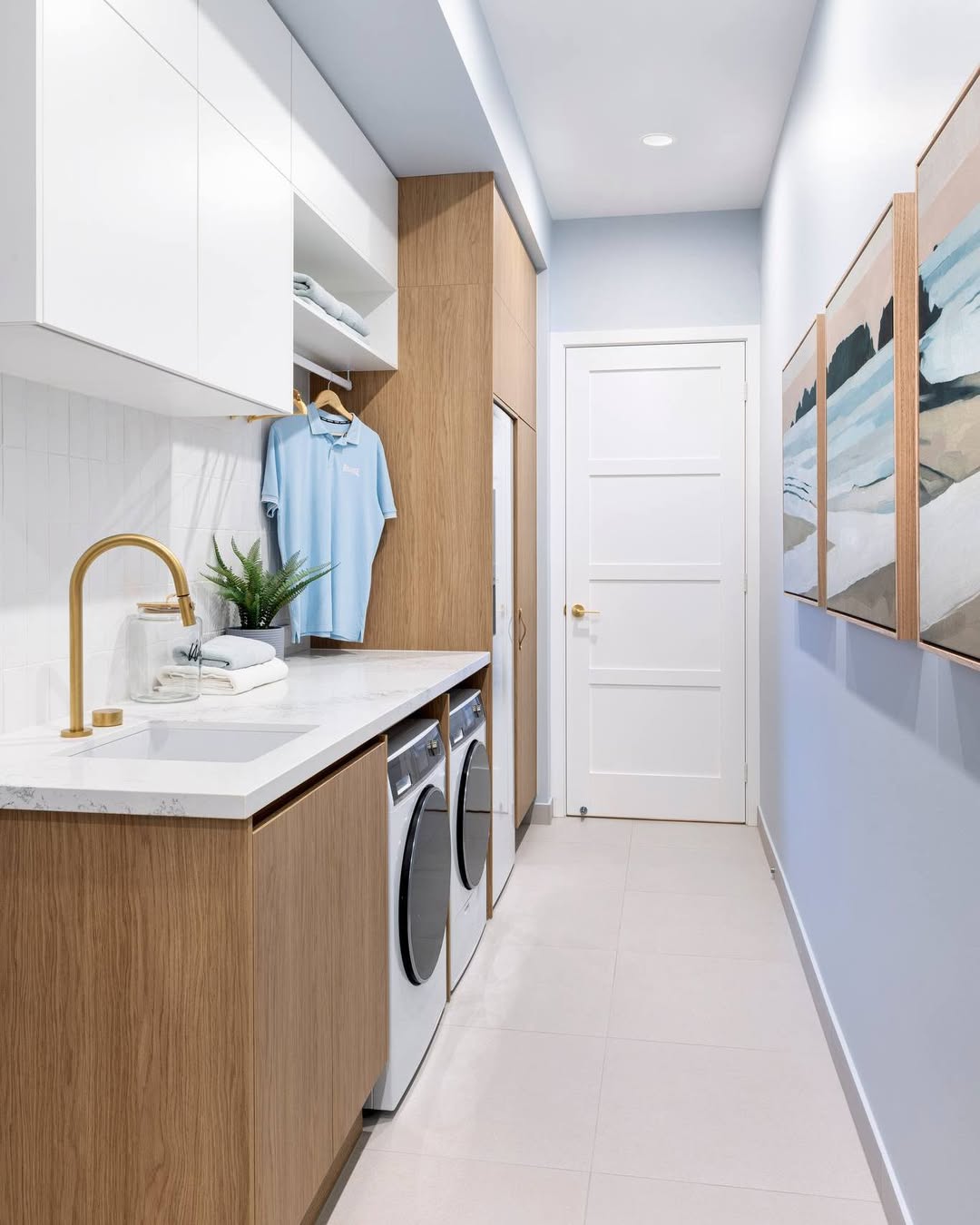 Modern laundry room with wood and white cabinets, washer and dryer, marble countertop, gold faucet, a hanging shirt, potted plant, framed artwork on a light blue wall—perfect for inspiring boho decor and fresh laundry room ideas.