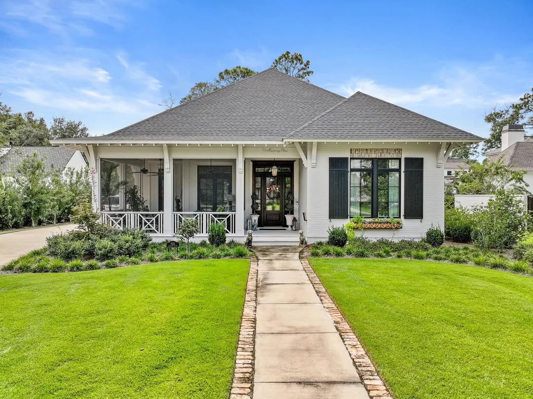 Single-story white house with gray roof, black shutters, front porch, flower box, manicured lawn, and a concrete walkway leading to the entrance under a blue sky—an inviting coastal cottage exterior perfect for design ideas 2025.