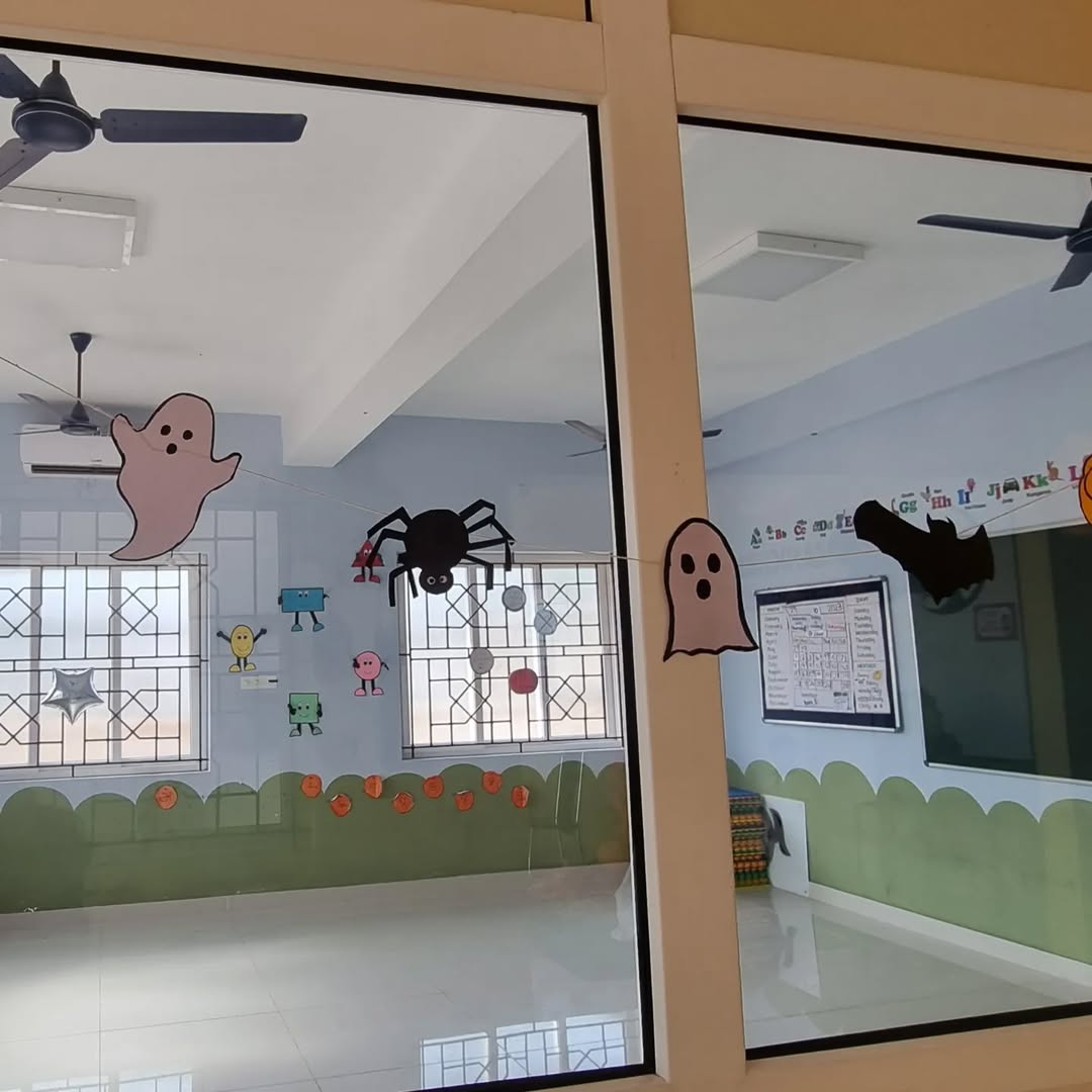 A classroom features creative Halloween window decoration ideas, with paper cutouts of ghosts, a bat, and a spider on glass windows, green wall decor, and children