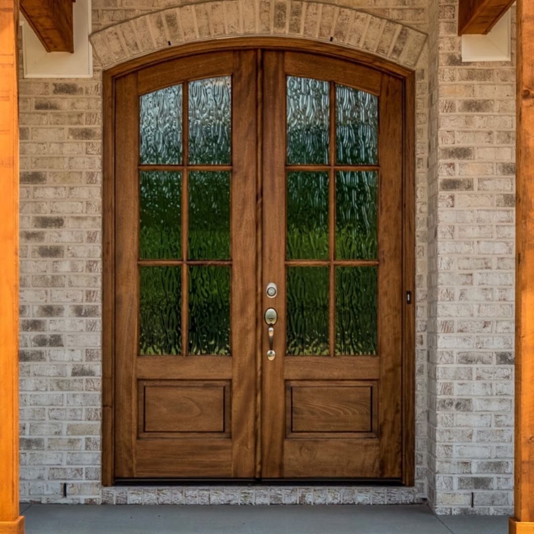 Double wooden front doors with glass windowpanes and textured glass create a welcoming double door entryway, set in a light brick exterior wall with an arched entry.