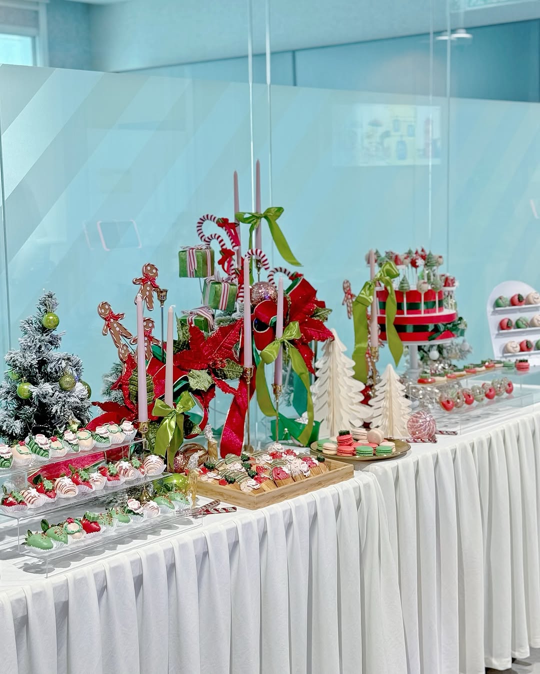 A decorated dessert table with pretty desserts, Christmas-themed treats, ornaments, candles, artificial trees, and festive ribbons on a white tablecloth—perfect for gathering dessert table ideas.