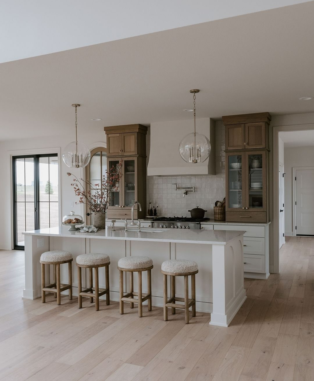 Modern kitchen design with a large island, four cushioned stools, light wood flooring, glass pendant lights, and natural wood cabinets with glass doors—perfect for those seeking farmhouse kitchen ideas.