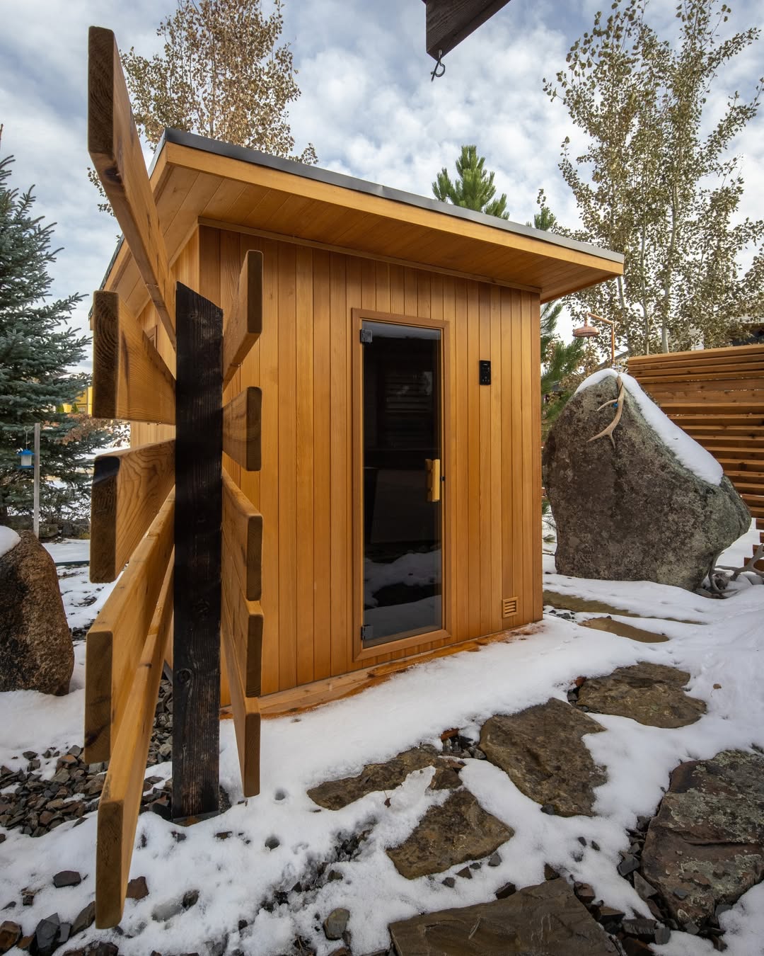 A small wooden outdoor sauna with a glass door sits on a stone path, surrounded by snow, rocks, and trees—one of the most gorgeous outdoor saunas for tranquil relaxation.