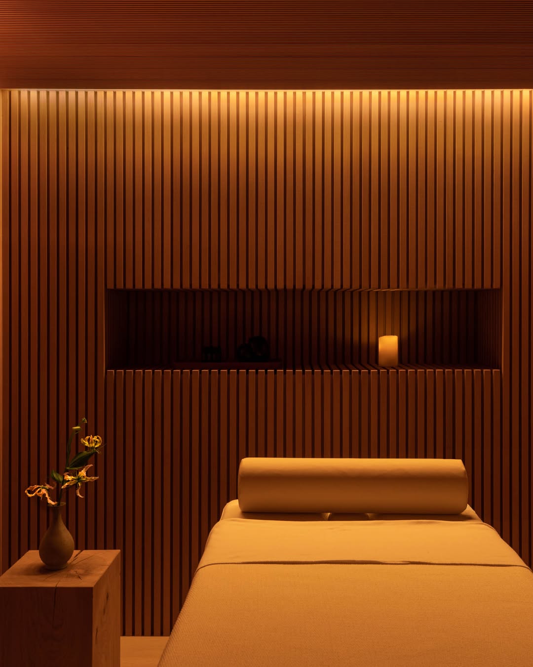 A massage bed with a cylindrical pillow sits in a warmly lit massage room featuring wooden slatted walls, a small vase with flowers, and a lit candle—perfect for inspiring new massage room ideas.