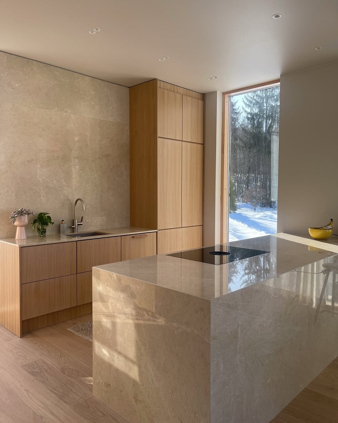 Modern oak kitchen with light wood cabinets, beige marble countertops, a sink, an induction cooktop, and a large window showing a snowy outdoor scene—perfect for those seeking inspiring oak kitchen ideas.