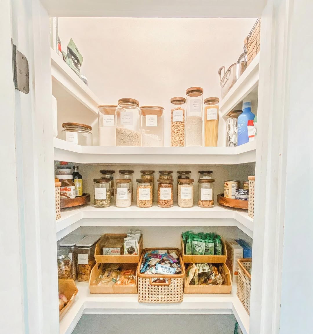 A neatly organized pantry with labeled containers, baskets, and jars showcasing pantry organization, holding various dry goods, snacks, and staples on white shelves.