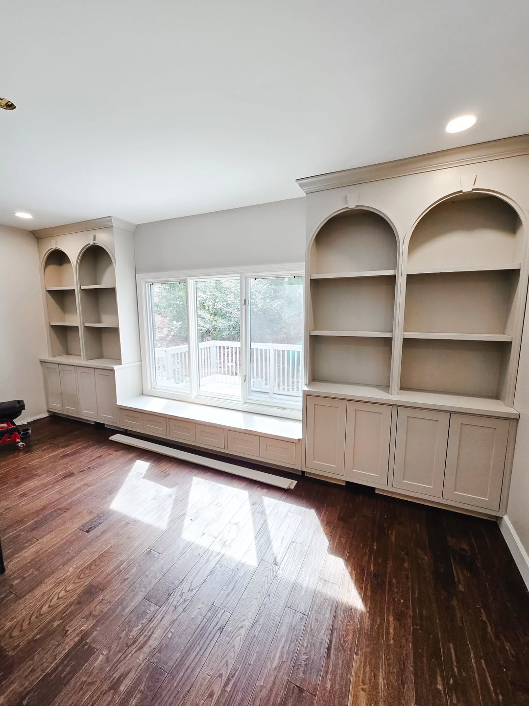 Built-in shelving units with arched tops flank a large window seat with storage drawers underneath, offering smart built-in storage and home organization against dark wood floors and crisp white walls.