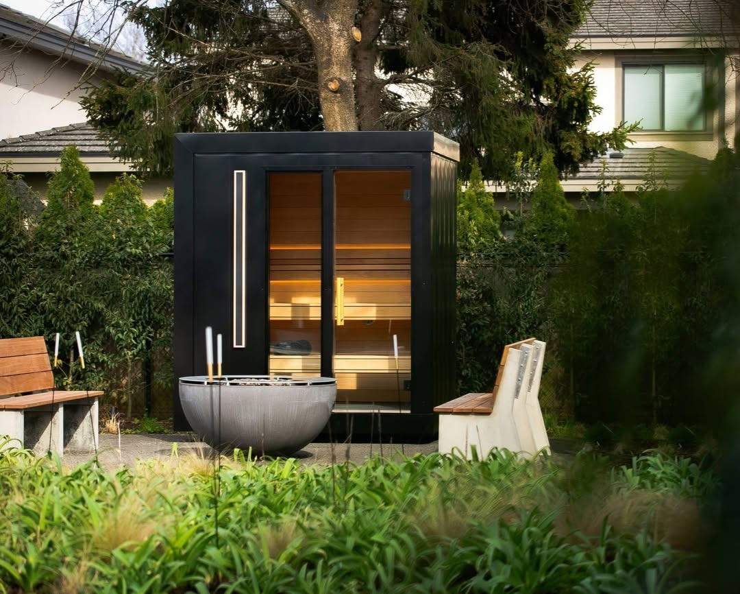 A modern black sauna with glass doors sits in a garden, surrounded by benches and greenery, with a stone fire pit in front—an inspiring example of gorgeous outdoor saunas.