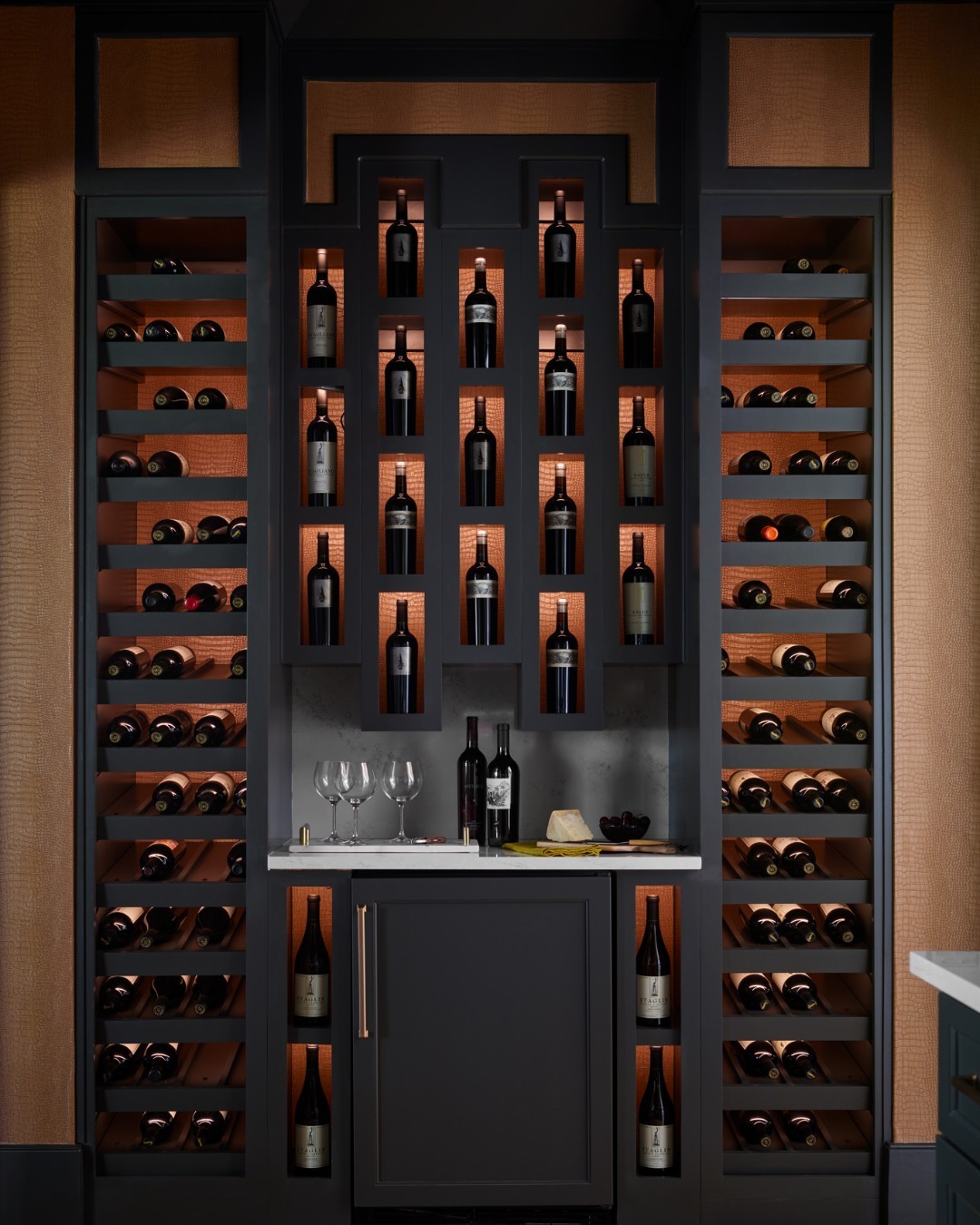 Modern wine cellar with dark wine storage shelves displaying multiple bottles, a small countertop with two glasses, three bottles, and a cheese plate, plus a mini fridge below—perfect inspiration for stylish wine rack ideas.