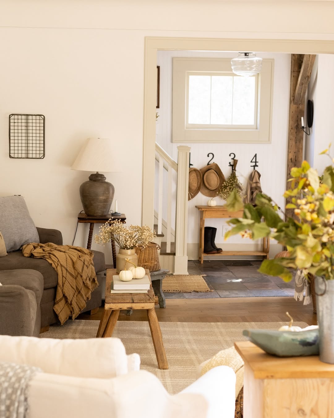 A cozy living room with neutral fall decor, a gray sofa, wooden side table, and a view into an entryway where hats hang on hooks labeled 1, 2, 3, and 4.