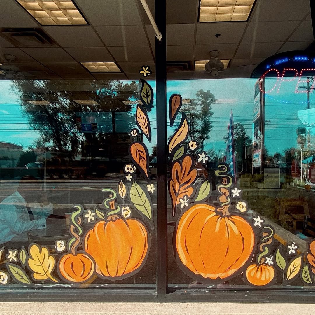 Storefront window decorated with painted orange pumpkins, green vines, and autumn leaves; a neon