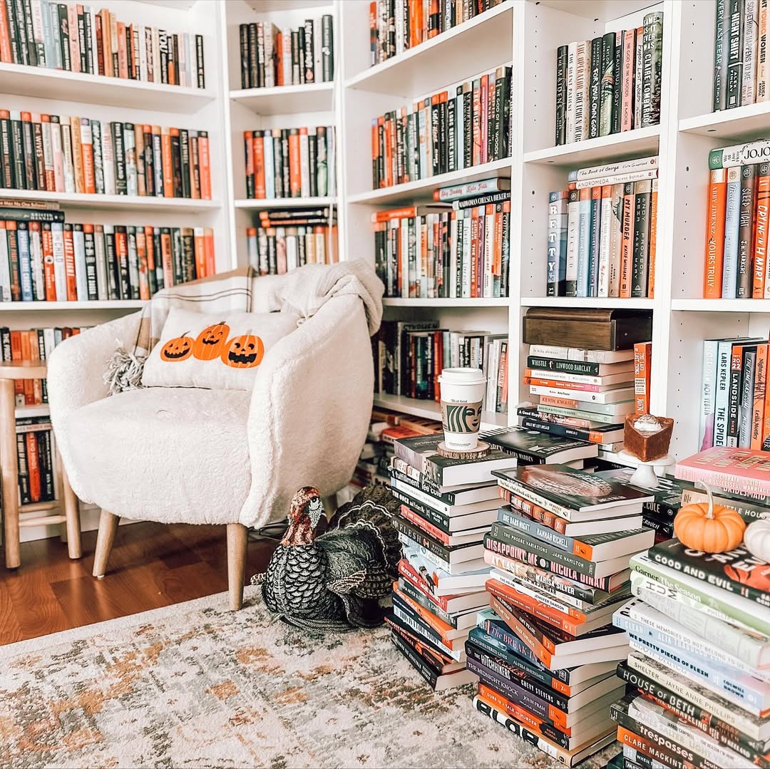 A cozy reading nook with a plush chair, pumpkin cushions, and piles of books, surrounded by white bookshelves—perfect inspiration for those seeking Cozy Book Nooks or unique Book Nook Ideas.