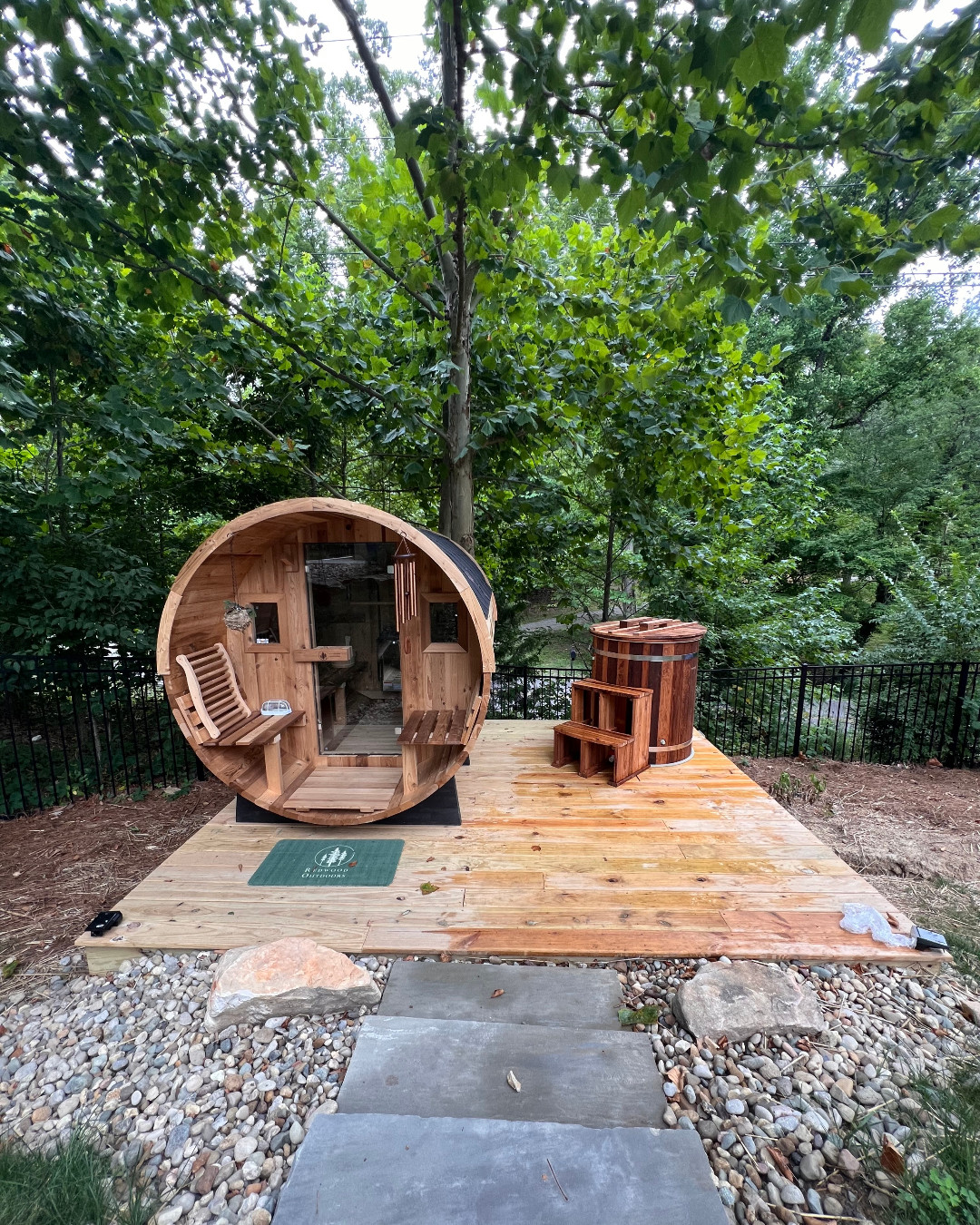 A round wooden sauna and hot tub are set on a wooden platform outdoors, surrounded by trees and greenery—gorgeous outdoor saunas like this offer inspiring outdoor sauna ideas, with stone steps leading up to the inviting deck.