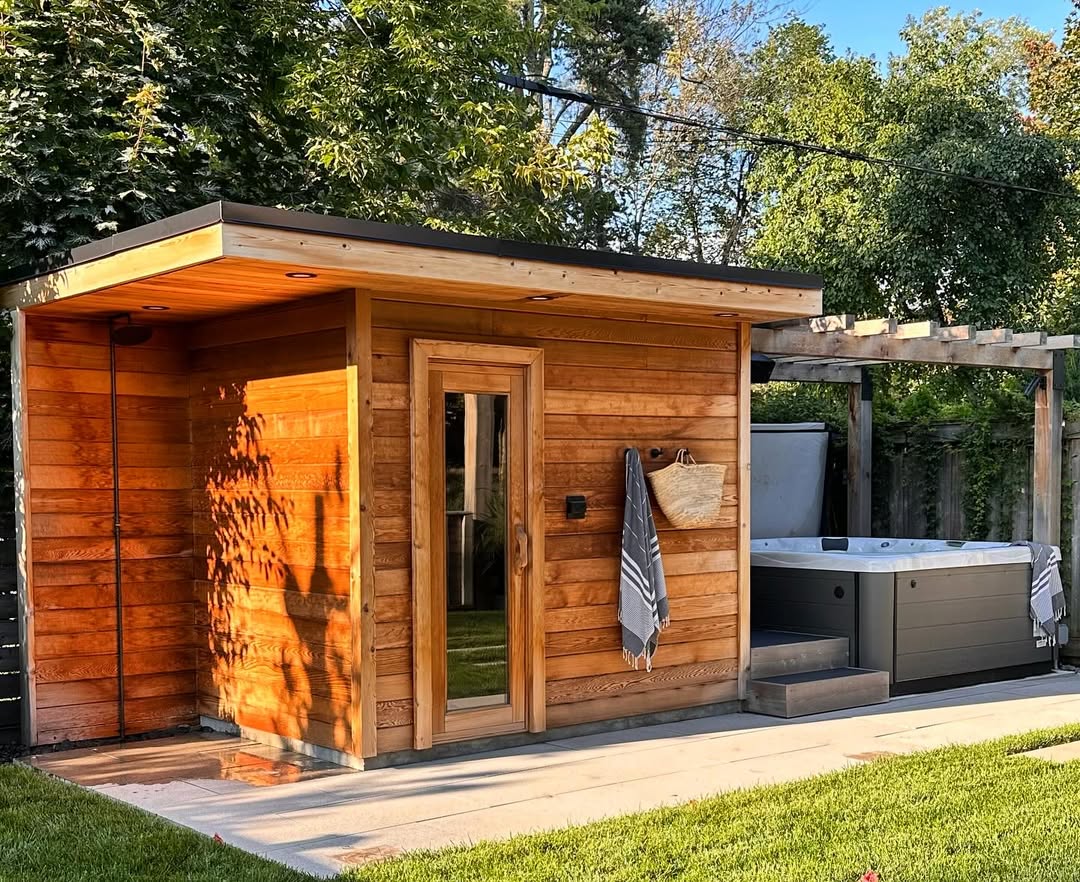 A modern wooden sauna with a glass door showcases stunning sauna design as it stands next to a gray hot tub under a pergola in a backyard with green grass and trees.
