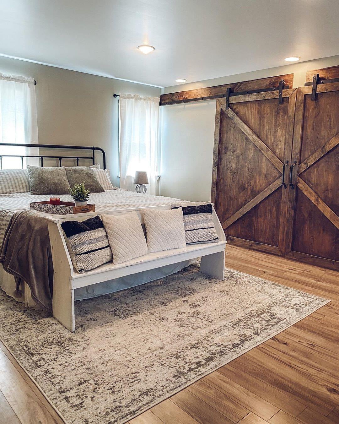 A bedroom with a neatly made bed, a bench with cushions at the foot, light wood flooring, large wooden sliding barn doors, and light walls with two windows—perfect inspiration for rustic bedroom ideas.
