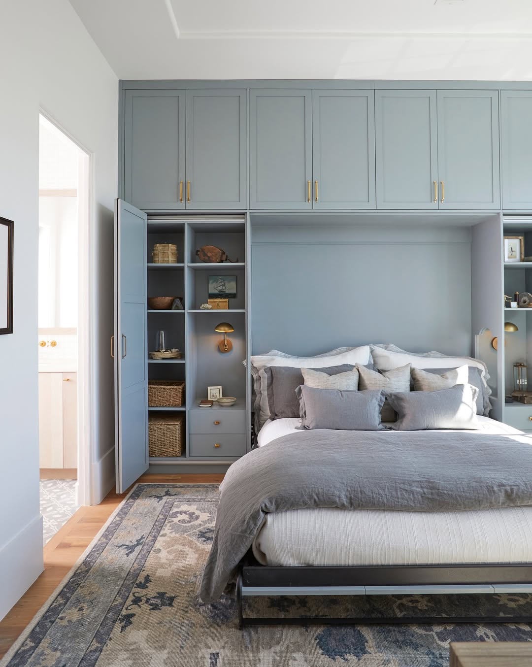 A bedroom with blue built-in storage cabinets above and around a bed, gray bedding, multiple pillows, open shelves with decor, and a patterned rug on a wooden floor—perfect for stylish home organization.