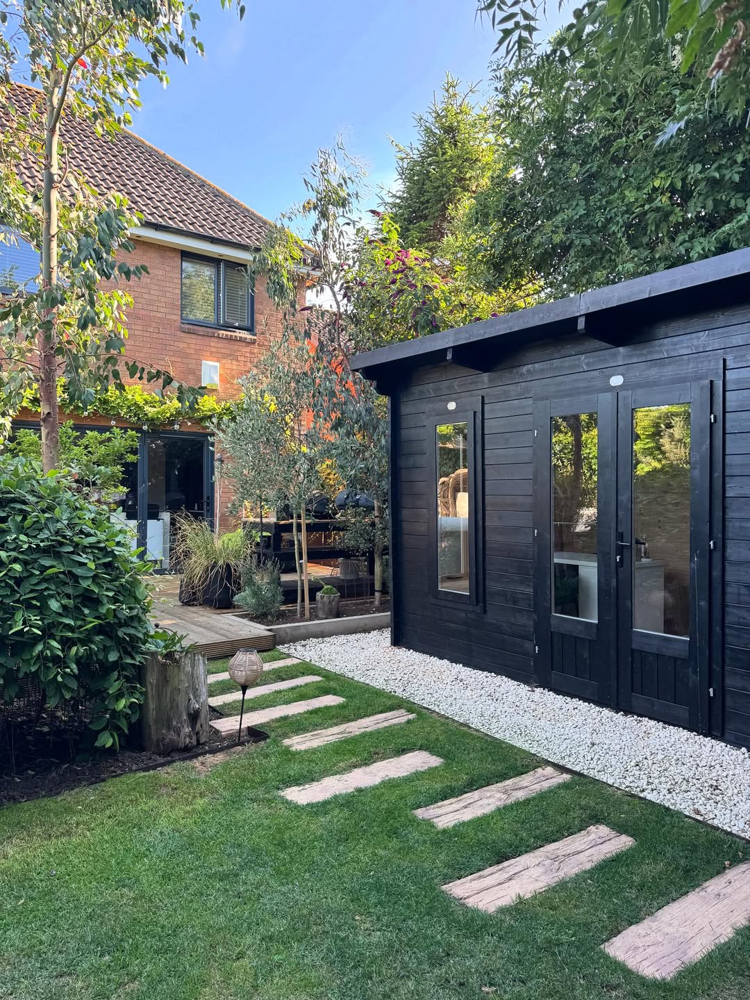 A modern backyard with a black garden shed, stone path, green grass, shrubs, and a view of a brick house with large windows—perfect inspiration for fresh Garden Ideas 2025.