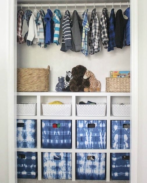 A neatly organized closet with children’s clothes hanging above shelves holding baskets, toys, and blue tie-dye storage bins—a perfect example of clever closet organization.