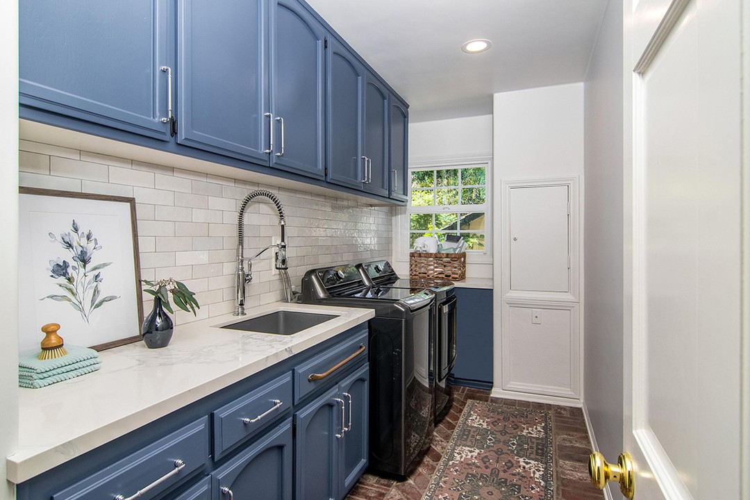 Laundry room with blue Laundry Room Cabinets, white countertops, farmhouse sink, black washer and dryer, white subway tile backsplash, framed art, and a window letting in natural light—perfect for those seeking Laundry Room Cabinet Ideas.