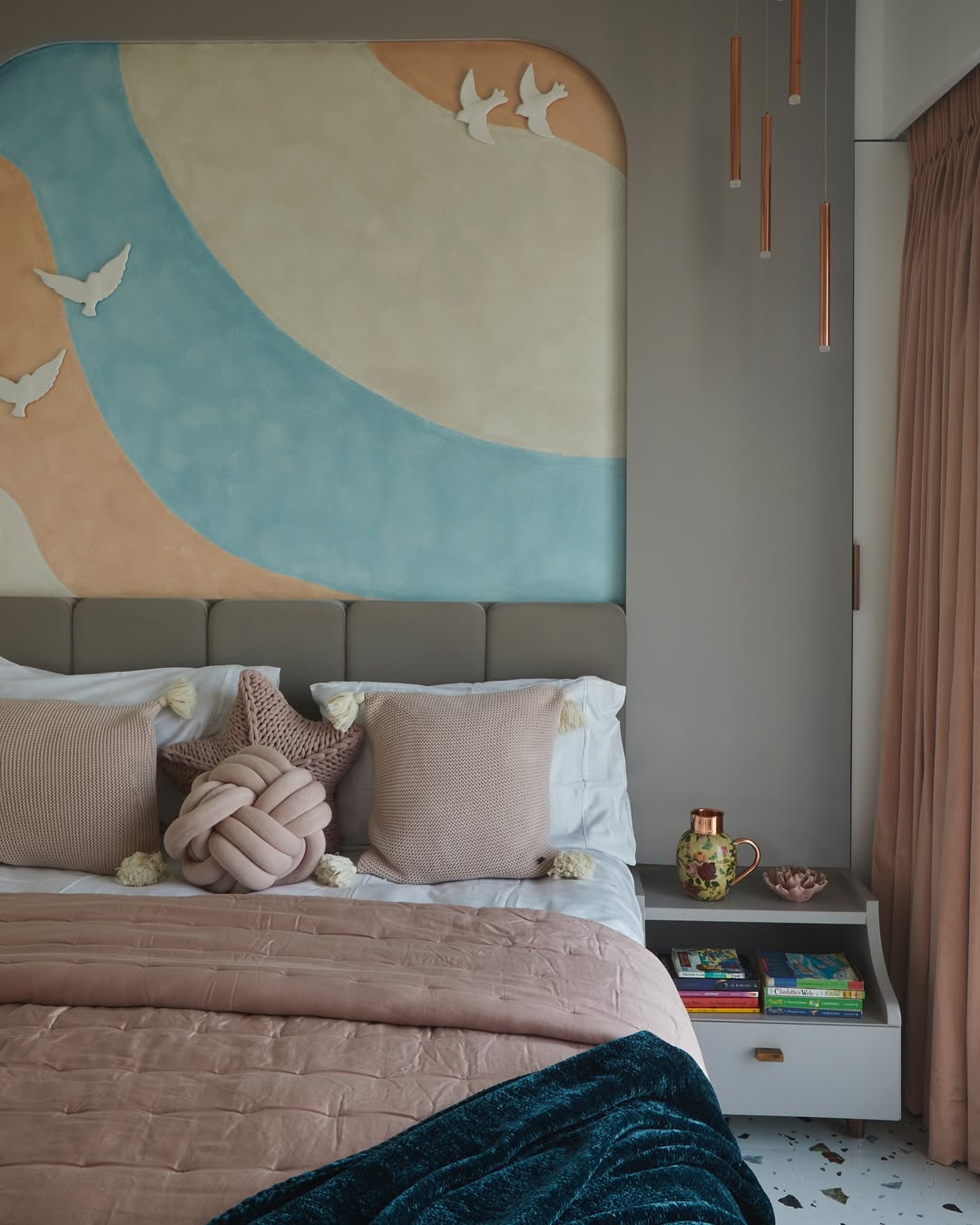A modern bedroom inspired by 2025 interior design trends features pastel bedding, plush pillows, a blue throw, a wall mural with flying birds, and a bedside table with books, a mug, and a vase—perfect for exploring new bedroom color schemes.
