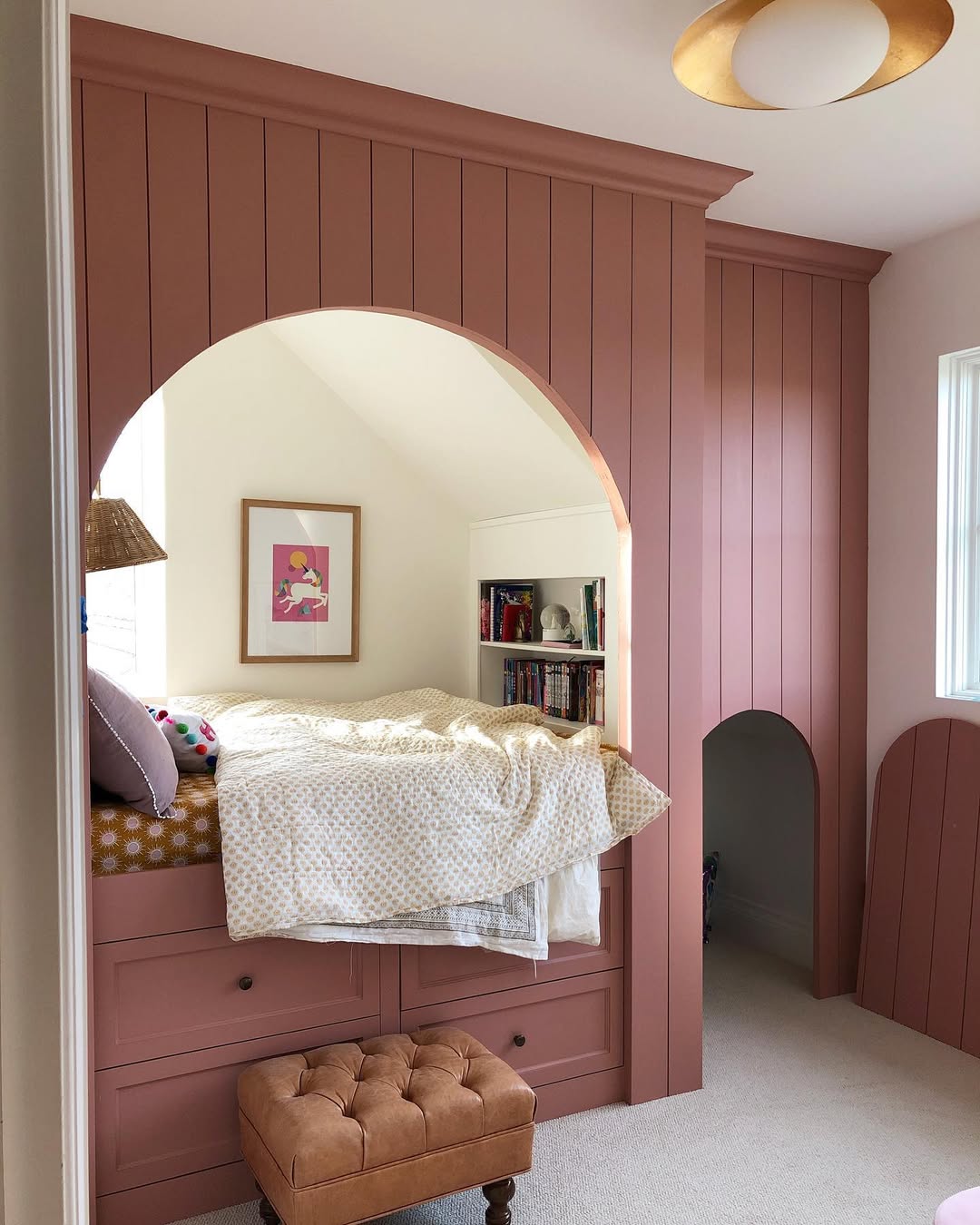 A cozy built-in pink alcove bed with drawers underneath, an arched entrance, a small bookshelf, framed art, and a tufted ottoman offers charming Pet Bedroom Ideas in this bright room.