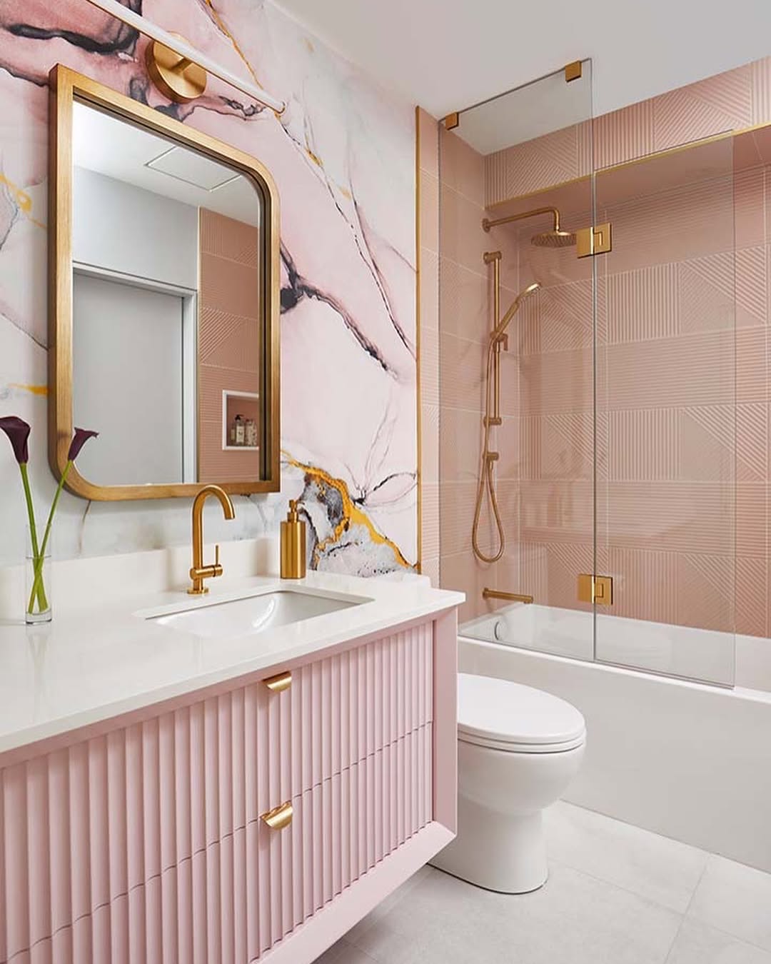 Modern bathroom with blush pink and gold accents, marble-patterned wall, glass shower enclosure, white countertop, pink vanity, gold fixtures, and a large rectangular mirror—perfectly complementing a sage green and blush pink bedroom.