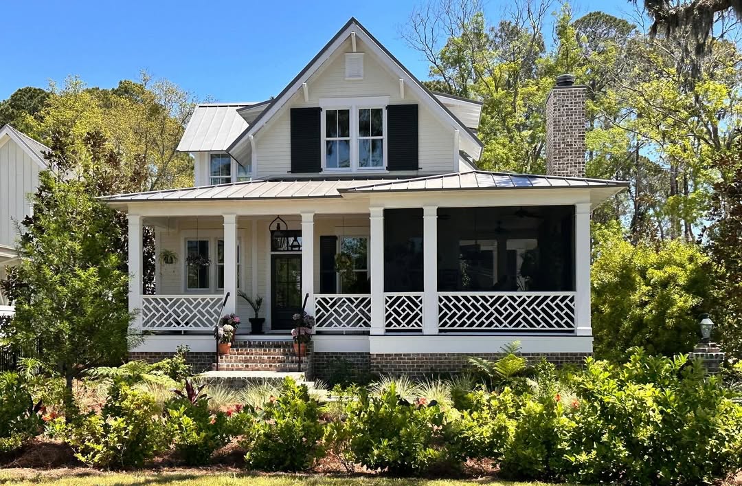 A white two-story house with a coastal cottage exterior design features a screened porch, black shutters, brick steps, and a landscaped front yard with shrubs and trees under a clear blue sky.