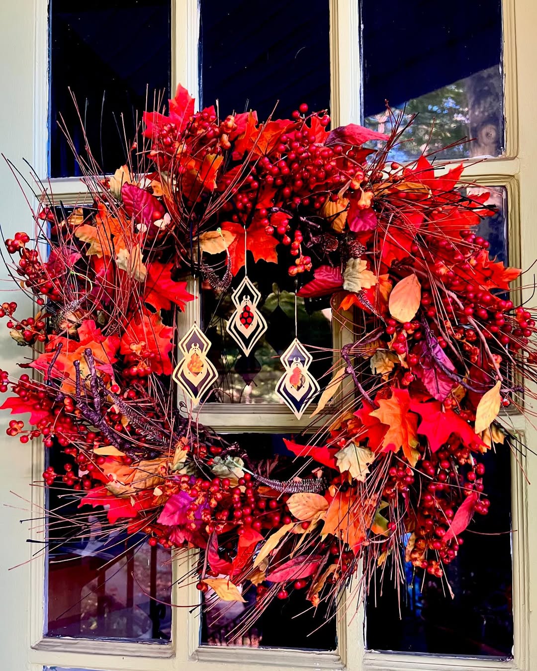 A decorative autumn wreath with red and orange leaves, berries, and twigs hangs on a glass-paneled door, featuring three geometric ornaments in the center—perfect inspiration for festive Halloween window decoration ideas.