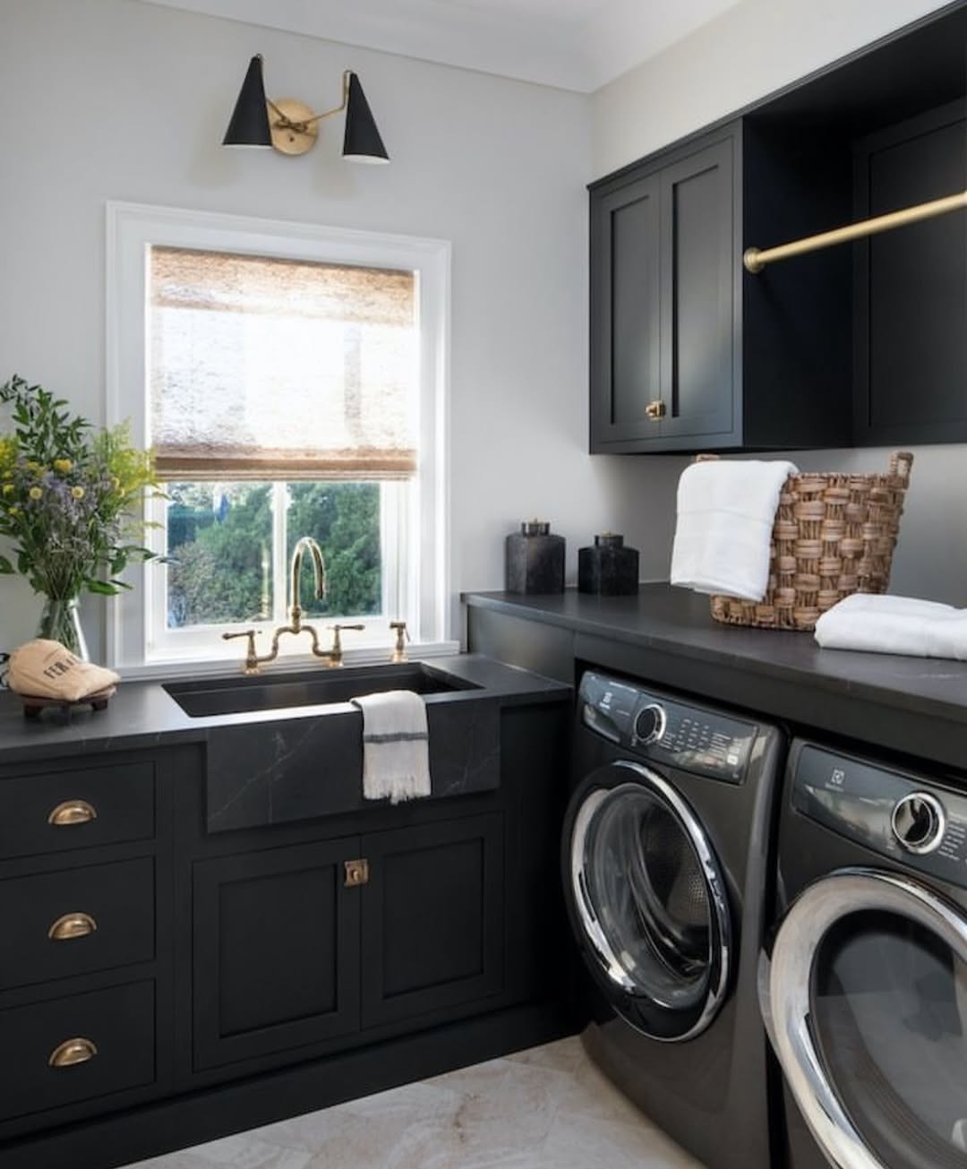 Modern laundry room with black laundry room cabinets, brass fixtures, a sink under a window, a washing machine, dryer, wicker basket, and a countertop for folding laundry—perfect inspiration for smart laundry room storage.