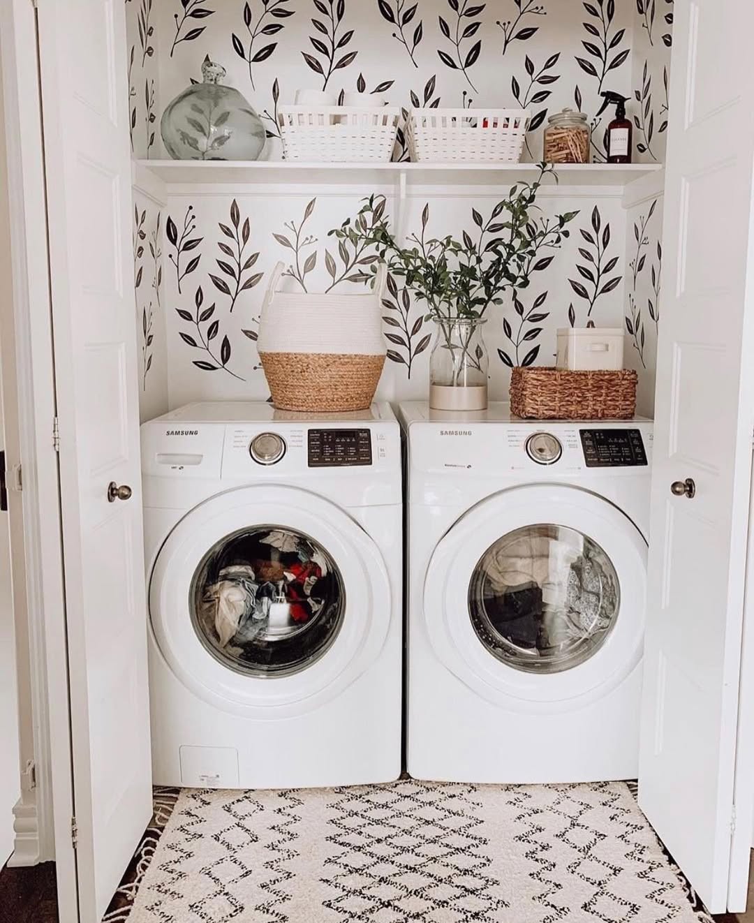 A laundry closet with a front-loading washer and dryer, shelves above holding baskets, jars, and a potted plant, plus leaf-patterned wallpaper, offers fresh Boho laundry room inspiration for those seeking stylish laundry room ideas.