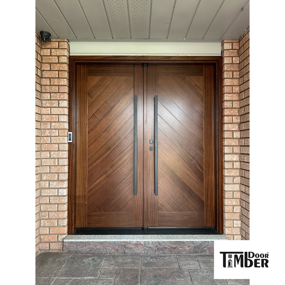A stunning double door entryway with a chevron pattern, set in a brick wall and featuring a tiled doorstep. The Timber Door logo is visible in the lower right corner, perfect for inspiring elegant entryway decor.