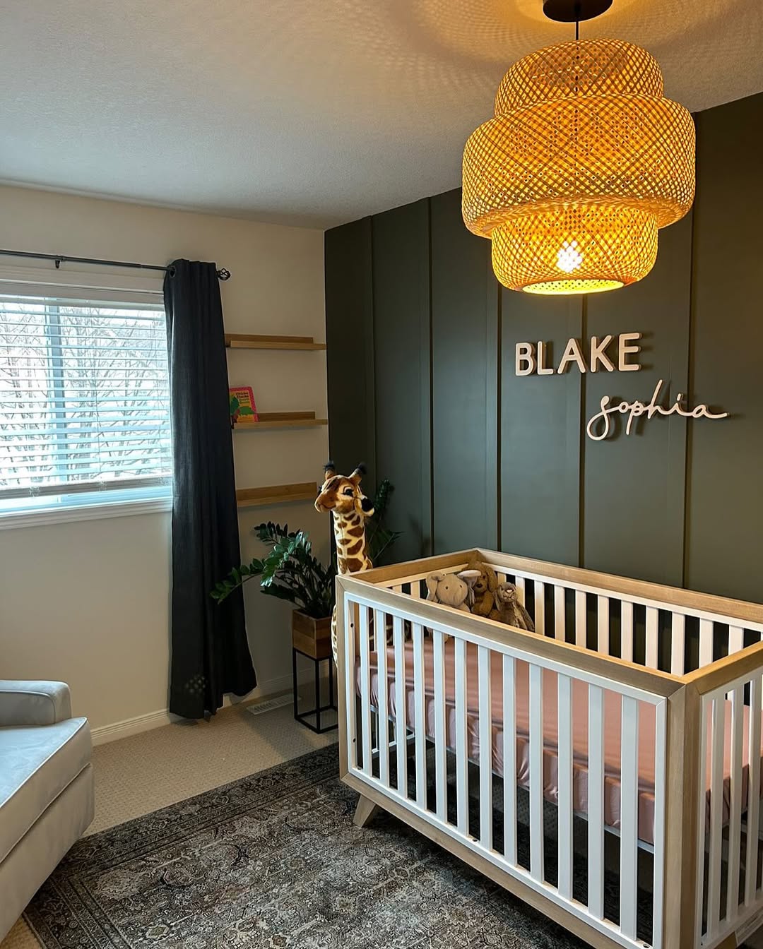 A nursery with a white crib, plush giraffe and lion toys, a large woven pendant light, dark green accent wall with