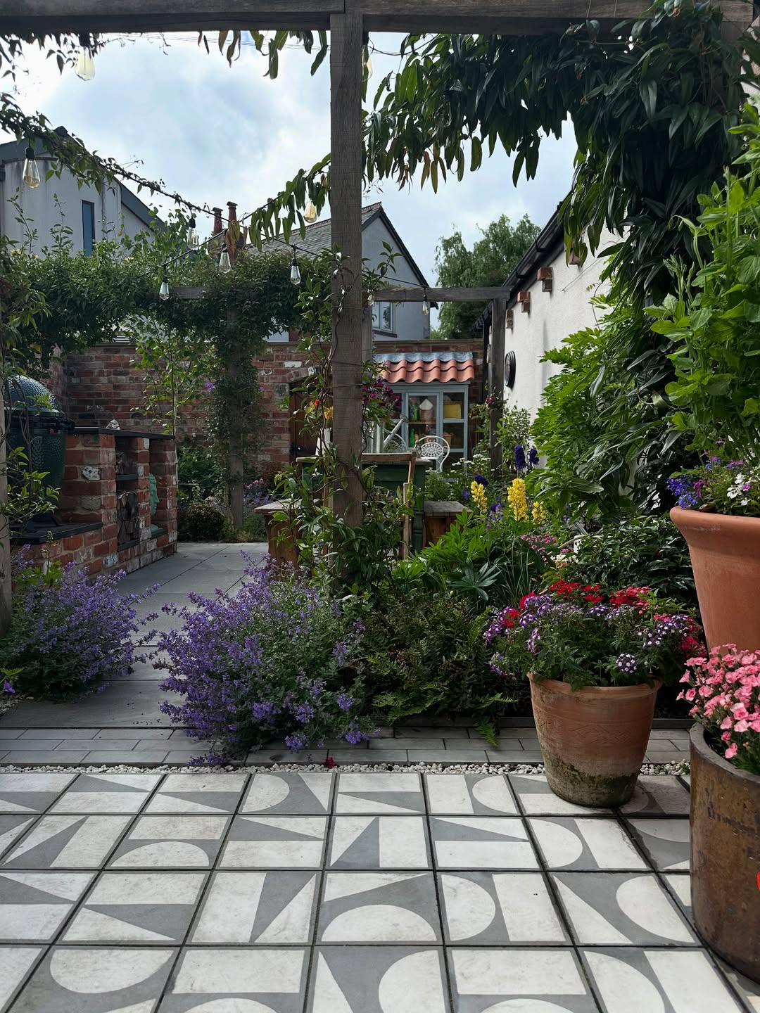 Outdoor patio with patterned tiles, potted flowers, and lush garden plants. A pergola with climbing vines frames a view of a small brick outbuilding—perfect inspiration for Garden Ideas 2025.