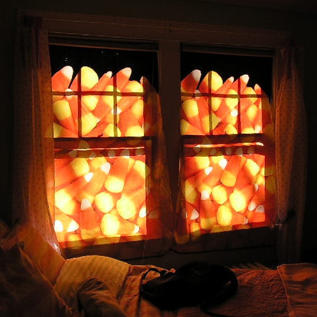 Two windows are covered with curtains featuring a large candy corn pattern, illuminated by light from behind—perfect Halloween window decoration ideas. A bed and pillows are visible in the foreground, completing the festive window decor.