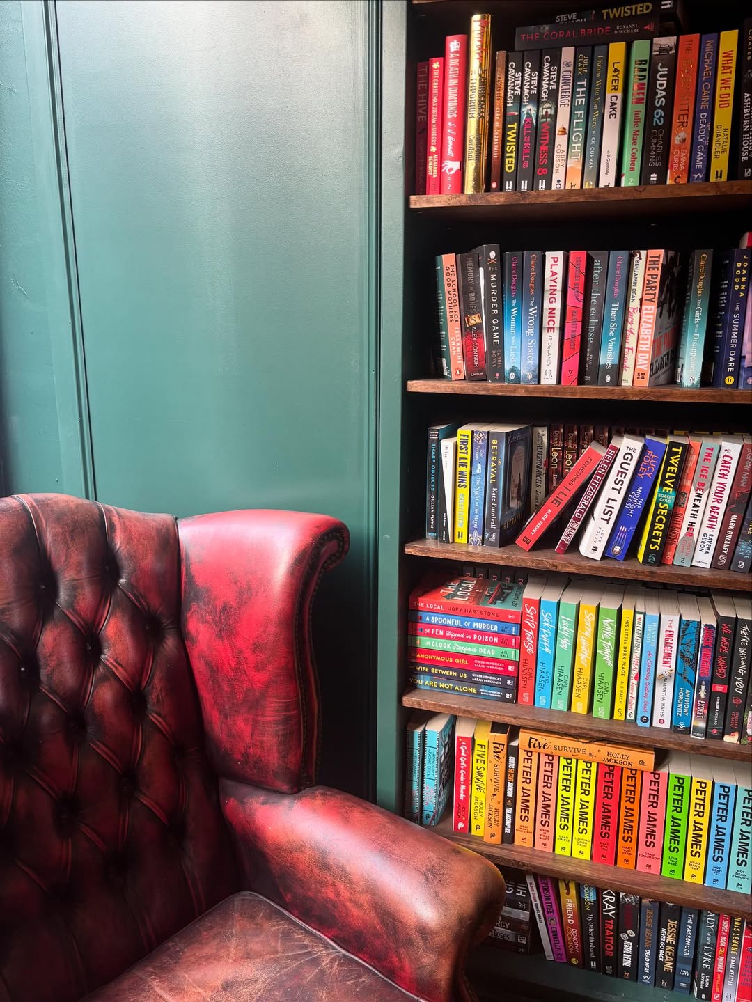 A worn red leather armchair sits next to a bookshelf filled with colorful, assorted books against a green wall, making it a perfect inspiration for cozy reading spaces and book nook ideas.