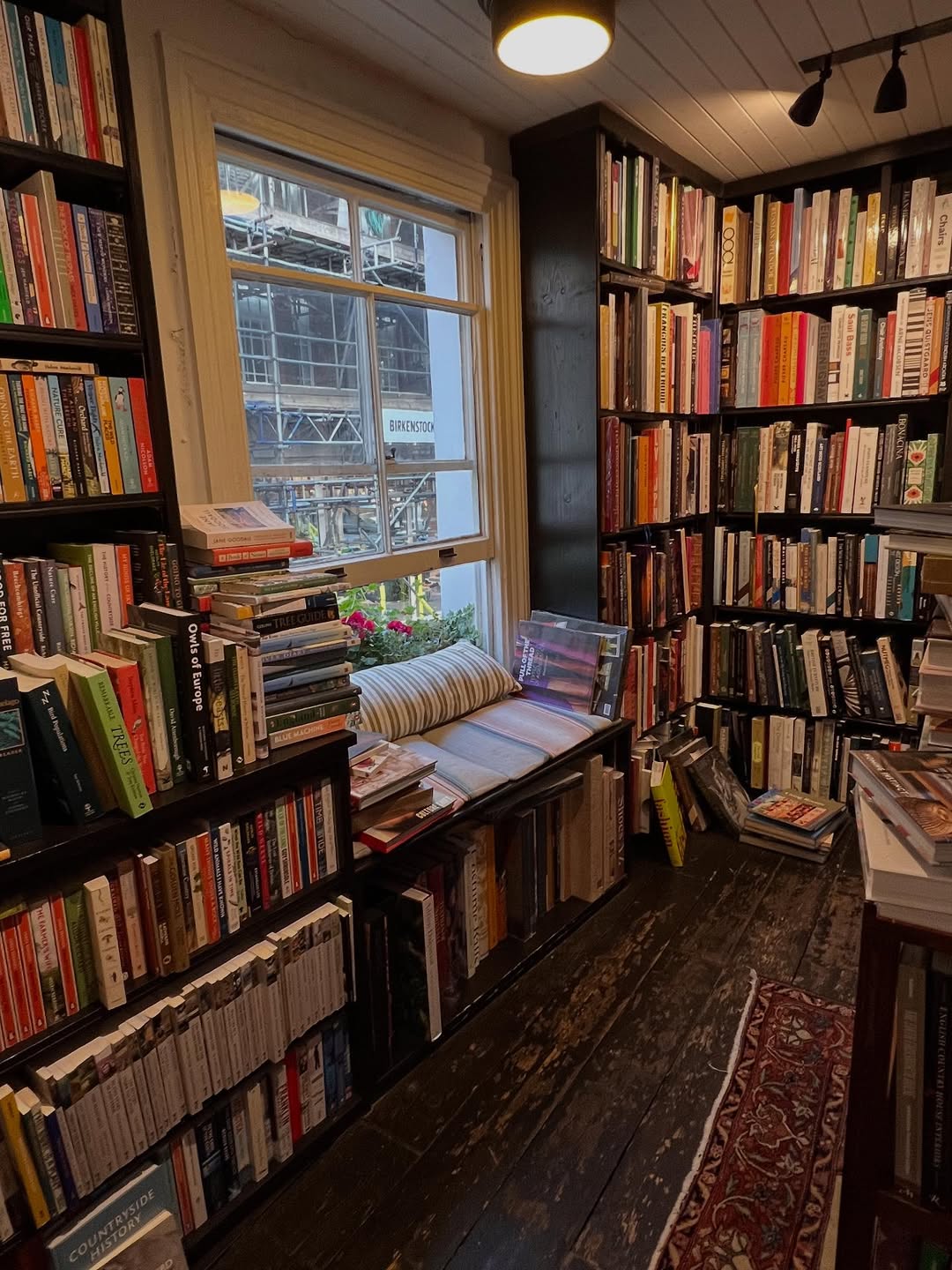A cozy reading nook in a bookshop corner with tall shelves filled with books, a wooden floor, a window seat with cushions, and a view of construction scaffolding outside—perfect inspiration for book nook ideas.