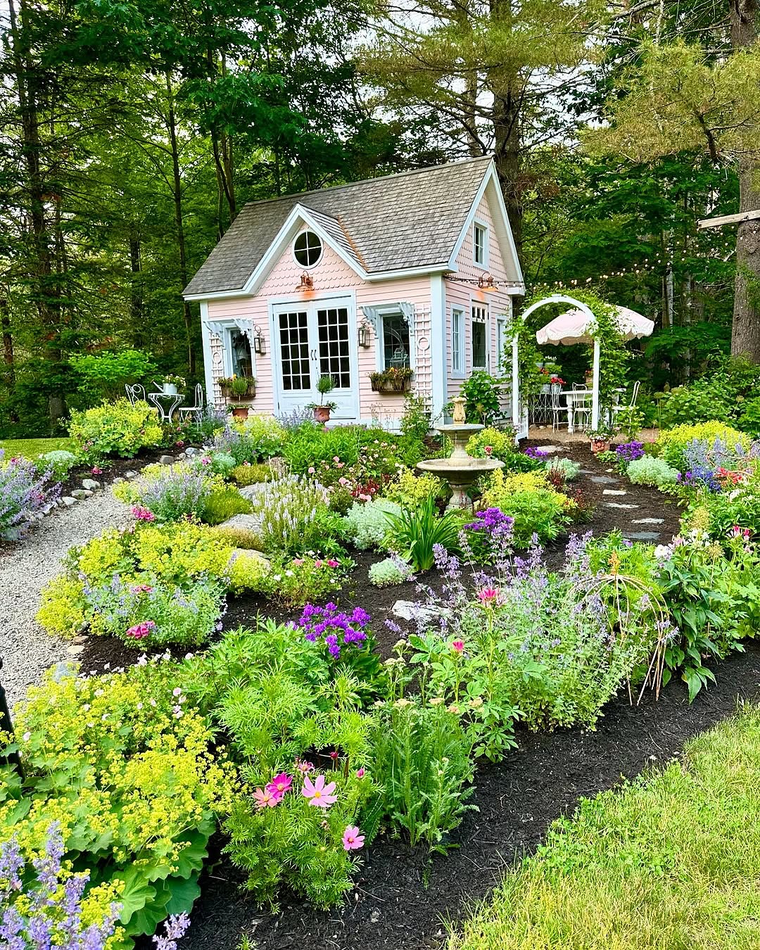 A small cottage sits amid an enchanting English cottage garden, bursting with blooming flowers, lush greenery, and a stone path, all surrounded by tall trees.