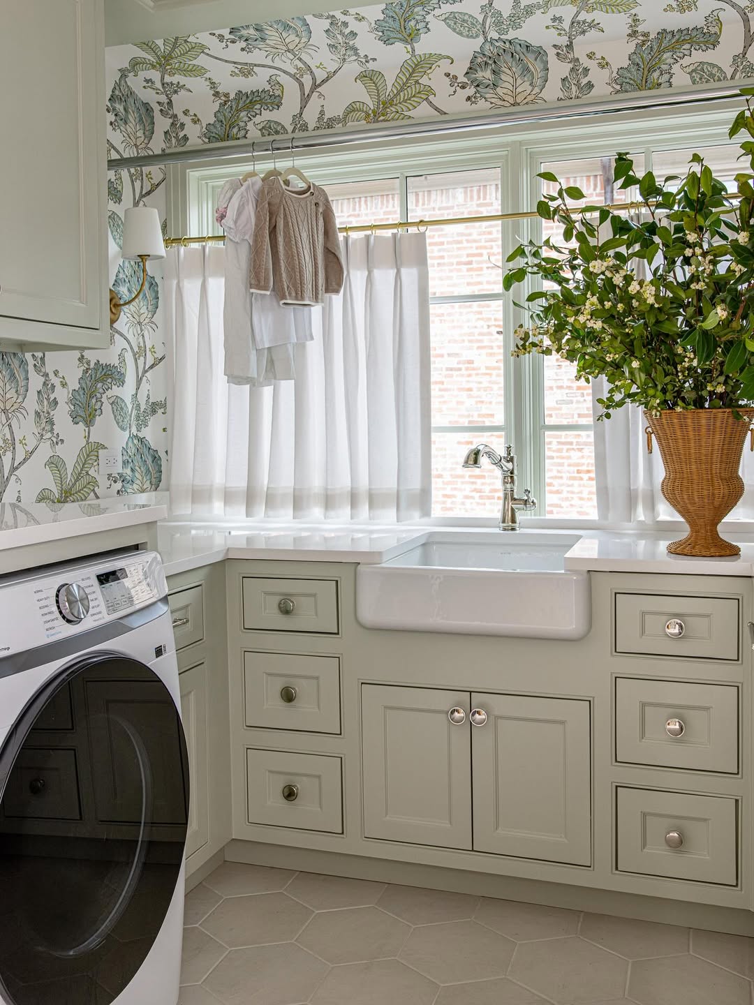 A laundry room design featuring a washing machine, farmhouse sink, light green cabinets, a window with sheer curtains, hanging clothes, and a vase with green branches—perfect inspiration for small laundry room spaces.