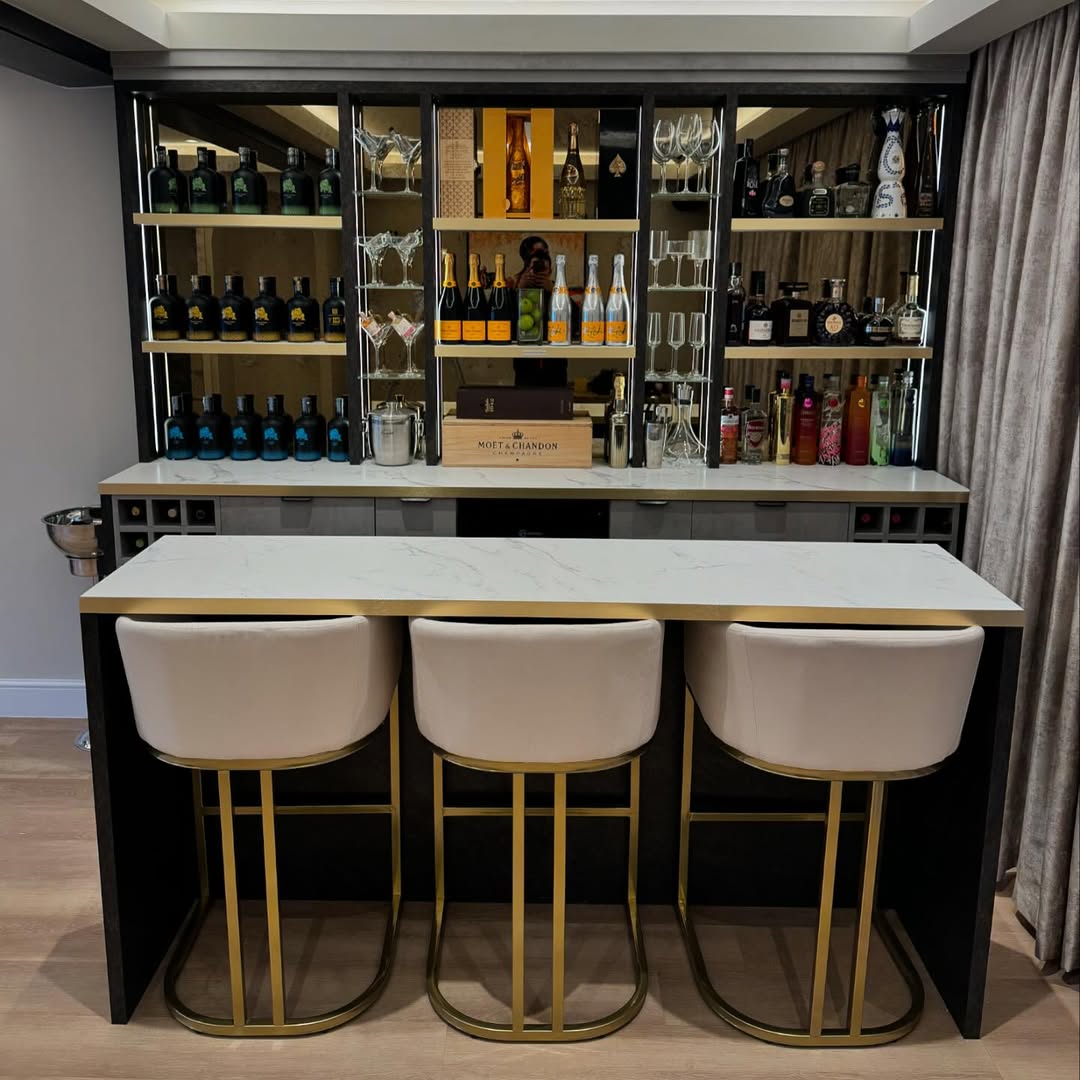 Home bar setup with three white and gold bar stools, a marble countertop, and shelves displaying various bottles of liquor, glassware, and a stylish wine rack for elegant wine storage.