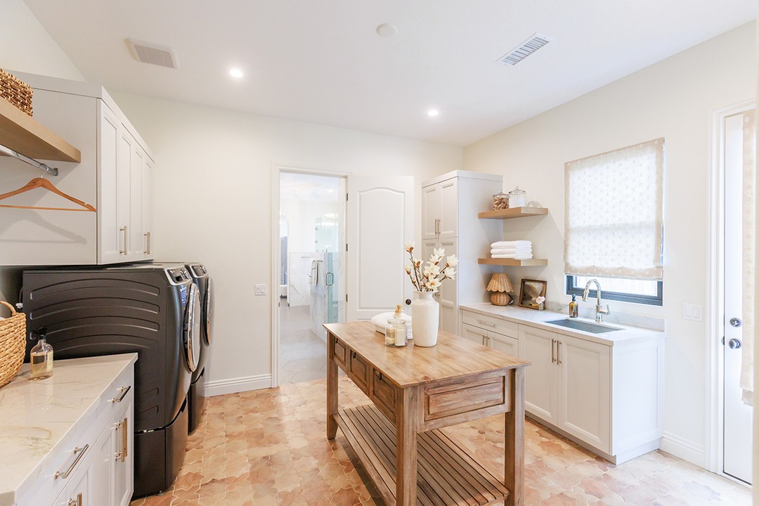 Bright laundry room with washing machine, dryer, sink, stylish Laundry Room Cabinet Ideas, wooden island table, shelves with towels, and a window with blinds.