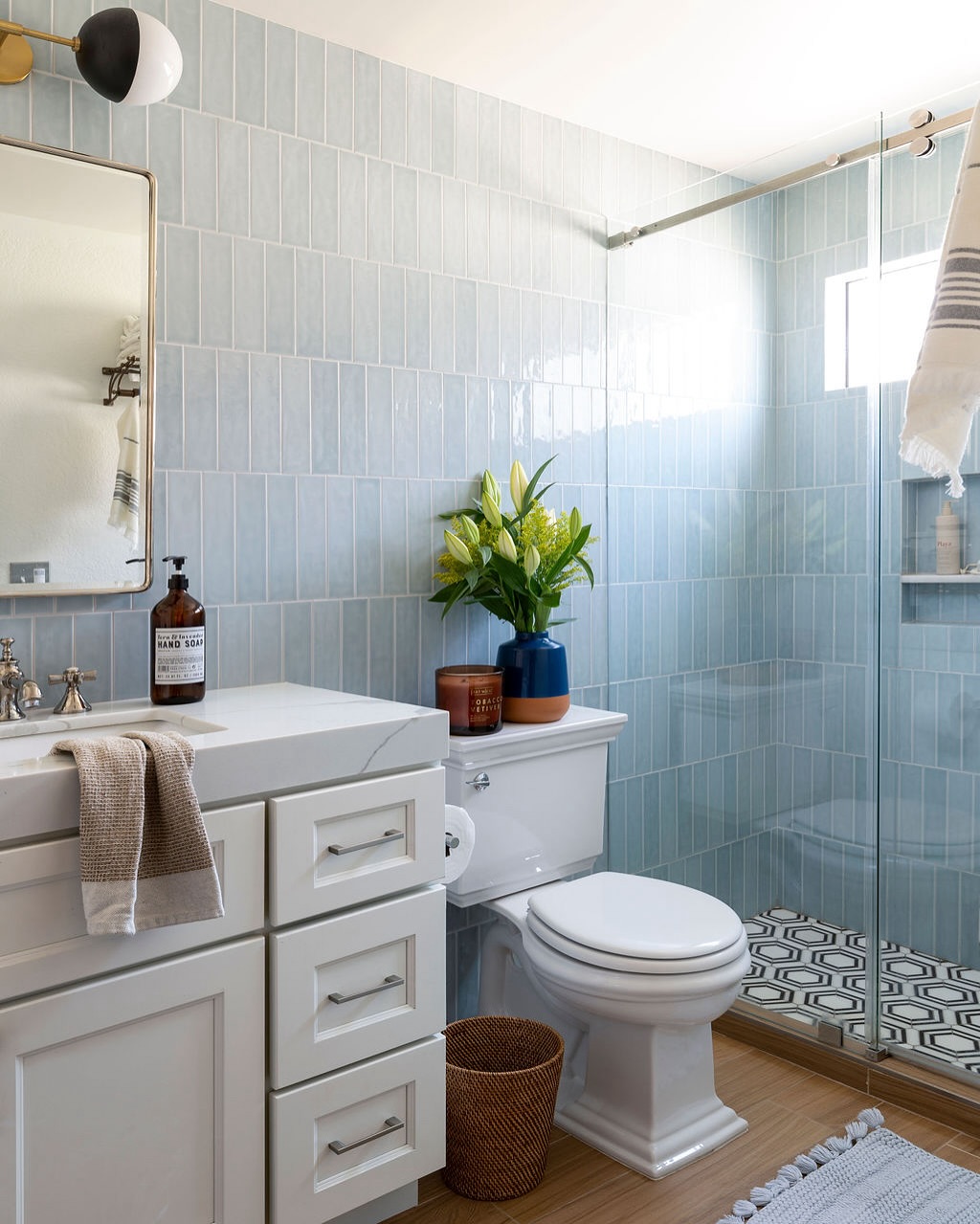A modern kids bathroom with light blue tiled walls, a white vanity, toilet, potted plants, a glass shower, and playful patterned floor tiles.