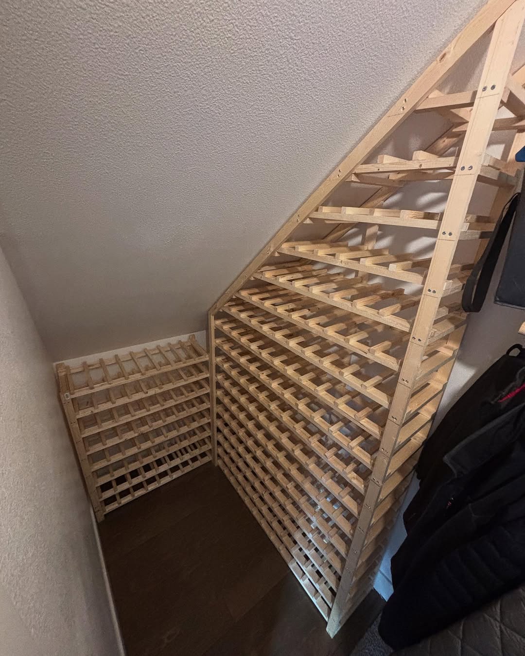 Wooden wine racks are installed under a sloped ceiling in a corner, maximizing storage space with multiple horizontal shelves for bottles—perfect for those seeking stylish under stairs storage or unique closet ideas.