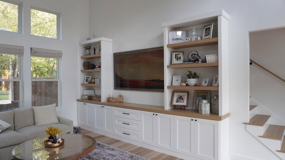Modern built-in shelves living room featuring white cabinets, a large TV, family photos, and decor. A staircase sits to the right while natural light streams in from windows on the left.