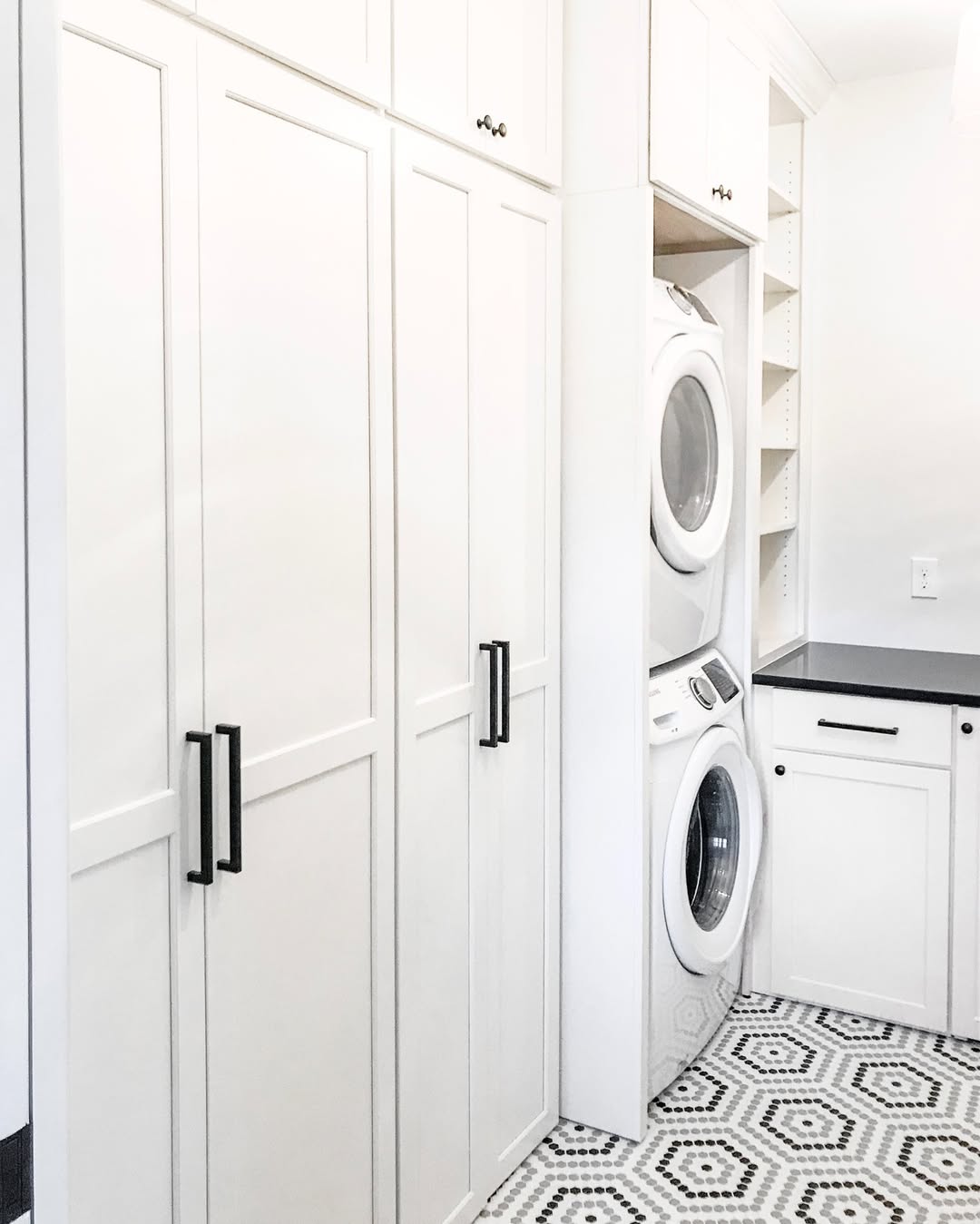 A stacked laundry room featuring white cabinets, a black countertop, and a patterned black-and-white tile floor for a clean, stylish look.